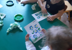 Trzy dziewczynki siedzą przy zielonym stoliku. Malują palcami niebieskie kwiaty z żółtym środkiem i zielonymi liśćmi.
