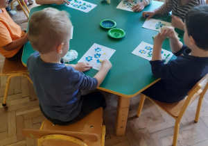 Przy zielonym stoliku siedzi pięciu chłopców i jedna dziewczynka. Malują palcami umoczonymi w farbie kwiaty o niebieskich płatkach, żółtym środku oraz zielonych liściach i trawie. Na środku stołu stoją dwa zielone talerzyki, na których znajduje się niebieska i zielona farba.