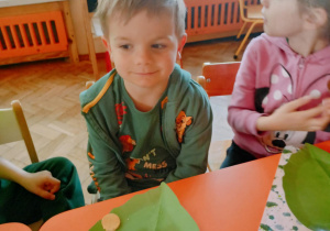 Na zdjęciu dwóch chłopiec i dziewczynka z grupy pomarańczowej, chłopcy siedzą przy stole i jedzą śnaidanie, dzieci ubrane są na zielono