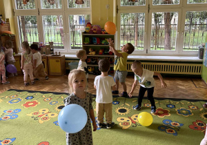 Na zdjęciu maluchy bawiące się żółtymi i niebieskimi balonami na dywanie w sali