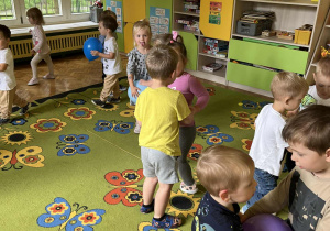 Na zdjęciu maluchy bawiące się żółtymi i niebieskimi balonami na dywanie w sali