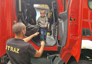 Na zdjęciu chłopiec z maluchów jest w wozie strażackim, obok wozu jest strażak