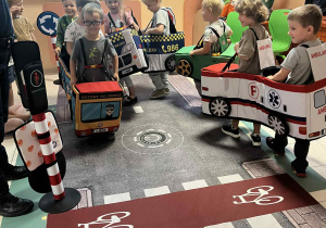 Na zdjęciach dzieci z grupy czerwonej podczas zajęć ze Strażą Miejską dzieci jeżdżą w samochodzikach nakładanych na dzieci, poruszają się za pomocą nóg