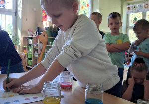 Na zdjęciu chłopczyk uzupełniający kartę pracy obok chłopca są kubeczki z różnymi kolorami