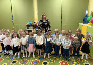 Na zdjęciu grupa żółta z wychowawcą dzieci są ubrane na galowo stoją na dywanie.