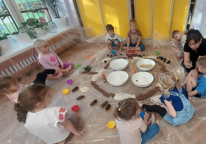 Na zdjęciu dzieci podczas zajęć sensoplastycznych dzieci siedzą w kole w środku koła są miseczki z różnymi materiałami