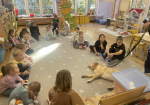 Na zdjęciu dzieci siedzą w kole , w środku koła leży pies labrador