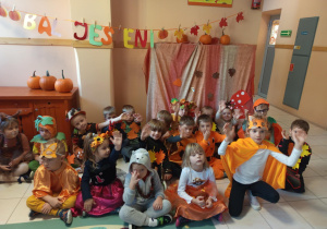 Na zdjęciu dzieci z grupy czerwonej dzieci siedzą na tle napisu Bal jesieni, dzieci są przebrane w nastroje inspirowane jesienią