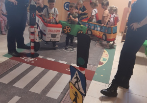 Ośmioro dzieci porusza się po macie przedstawiającej ulicę. Dzieci mają założone na siebie autochodziki: ambulansu, zielonego samochodu osobowego i autobusu. Po bokach rozłożonej maty stoją Panie ze straży miejskiej.