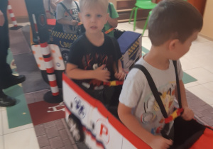 Sześcioro dzieci porusza się po macie przedstawiającej ulicę. Dzieci mają założone na siebie autochodziki: ambulansu, samochodu straży miejskiej.