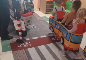 Siedmioro dzieci porusza się po macie przedstawiającej ulicę. Dzieci mają założone na siebie autochodziki: ambulansu, samochodu straży miejskiej i autobusu.