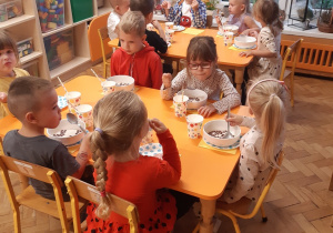 Sześć dziewczynek i pięciu chłopców siedzi przy dwóch żółtych stołach. Dzieci jedzą śniadanie na zastawie w kolorowe kropki.