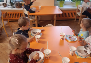 Sześioro dzieci siedzi przy stolikach. Dzieci jedzą śniadanie na zastawie w kolorowe kropki.