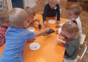 Trzy dziewczynki i trzech chłopców siedzi przy żółtym stoliku. Dzieci malują farbami kropki na białych, papierowych talerzykach.