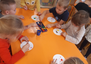 Cztery dziewczynki i dwóch chłopców przy żółtym stoliku maluje farbami kropki na białych, papierowych talerzykach.