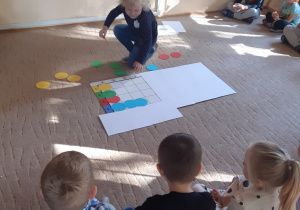 Dzieci siedzą na beżowym dywanie. Na środku dywanu leży mata do kodowania, na której dziewczynka układa kolorowe, papierowe koła.