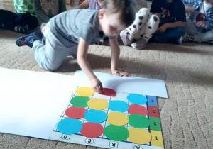 Dzieci siedzą na beżowym dywanie. Na środku dywanu leży mata do kodowania, na której chłopiec układa kolorowe koła.