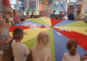 Dzieci ustawione są w kole, w rękach trzymają kolorową chustę animacyjną. Na chuście położone są foliowe, nadmuchane powietrzem woreczki.