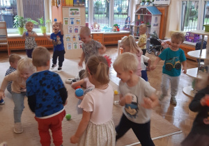 Dzieci ustawione są w rozsypce na beżowym dywanie i brązowej podłodze. Bawią się kolorowymi piłeczkami.