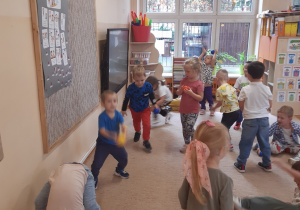 Dzieci ustawione są w rozsypce na beżowym dywanie i brązowej podłodze. Bawią się kolorowymi piłeczkami.