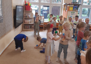 Dzieci ustawione są w parach na beżowym dywanie i brązowej podłodze. Bawią się kolorowymi piłeczkami.