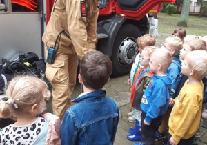 Dzieci stoją przed stażakiem, który pokazuje dzieciom młot pneumatyczny. W tle widać wóz strażacki.