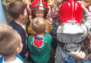 Dwaj chłopcy przymierzają hełmy strażackie, trzech chłopców przygląda się im. W tle widać osoby dorosłe.