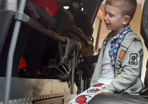 Chłopiec w szarej bluzie i szarych spodniach siedzi we wnętrzu strażackiego wozu.