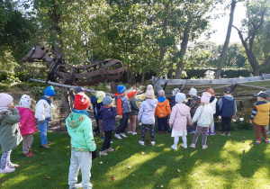 Dzieci stoją w rozsypce na zielonej trawie. W tle widać metalową rzeźbę konia i drewniany wóz.