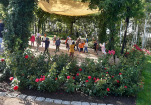 Dzieci stoją na betonowym okręgu. Nad dziećmi rozciągnięty jest żółty materiał. Z przodu zdjęcia widać krzaki czerwonych róż.