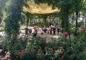 Dzieci stoją na betonowym okręgu. Nad dziećmi rozciągnięty jest żółty materiał. Z przodu zdjęcia widać krzaki czerwonych róż.