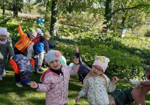 Dzieci stoją na zielonej trawie. W tle widać kolorową rzeźbę żaby oraz dwa ule.