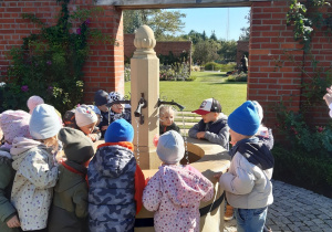 Dzieci ustawione są wokół beżowej fontanny. W tle widać mur z czerwonych cegieł.