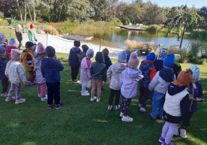 Dzieci stoją na zielonej trawie. W tle widać staw z pomostem i łódkę.