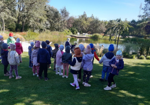 Dzieci stoją na zielonej trawie. W tle widać staw z pomostem, łódkę i drzewa.