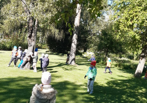 Dzieci stoją na zielonej trawie, niektóre przytulają się do drzewa.