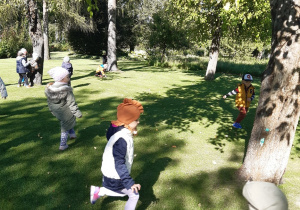 Dzieci biegają po zielonej trawie pomiędzy drzewami.