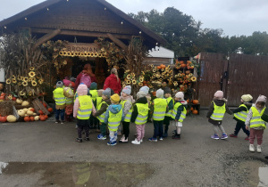 Dzieci ubrane w żółte, odblaskowe kamizelki stoją w parach. W tle widać wejście do Dobronianki ozdobione żółtymi i pomarańczowymi dyniami oraz żóltymi słonecznikami.