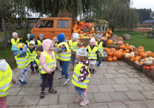 Dzieci ubrane w żółte, odblaskowe kamizelki stoją na tle pomarańczowego samochodu, na którym położone są dynie. Z boku na chodniku ułożone są skrzynki wypełnione dyniami.