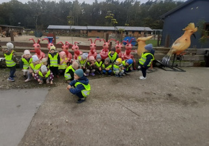 Dzieci ubrane w żółte, odblaskowe kamizelki kucają przed różowymi, plastikowymi figurami mrówek. Po prawej stronie zdjęcia stoją dwie figurki ptaków.