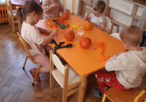 Dwie dziewczynki i dwóch chłopców siedzi przy pomarańczowym stoliku. Na stoliku leżą małe, pomarańczowe dynie, biały klej w plastikowym pojemniku, bibuła w kolorze żółtym, pomarańczowym, zielonym i czarnym.
