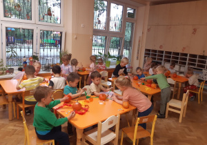 Dzieci siedzą przy pomarańczowych stolikach. Na stołach leżą pomarańczowe, małe dynie, biały klej w plastikowych pojemnikach, żółta, pomarańczowa i zielona bibuła oraz papierowe oczy i usta.