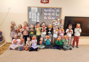 Dzieci pozują do zdjęcia na beżowym dywanie. Dzieci trzymają w rękach zrobione przez siebie dyniowe stworki.