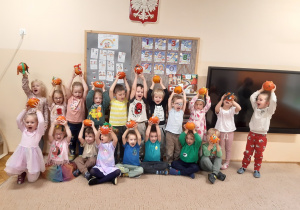 Dzieci pozują do zdjęcia na beżowym dywanie. Dzieci trzymają w uniesionych do góry rękach zrobione przez siebie dyniowe stworki.