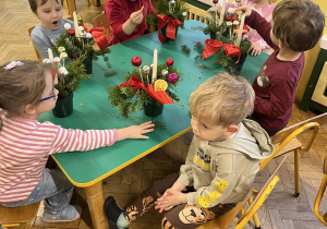 Na zdjęciu dzieci siedzące przy stole dzieci wykonują stroik świąteczny