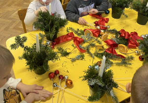 Na zdjęciu dzieci siedzące przy stole dzieci wykonują stroik świąteczny