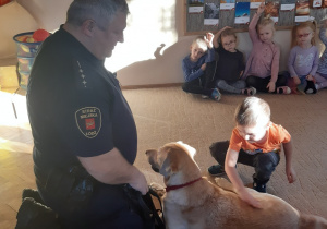 Na beżowym dywanie siedzą dzieci i strażnik miejski. Przed pracownikiem straży miejskiej leży beżowy pies, którego głaszcze chłopiec w pomarańczowej bluzce.