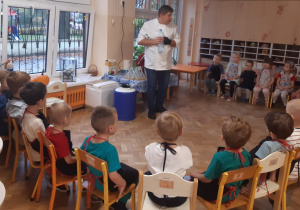 Dzieci siedzą na krzesłach ustawionych w półkole. Przed dziećmi stoi mężczyzna w białym fartuchu, który w rękach trzyma plastikową butelkę wypełnioną wodą.