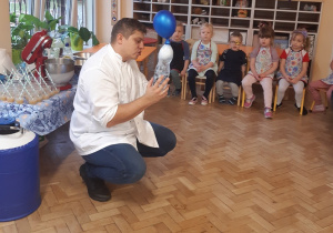Dzieci siedzą na krzesełkach i przyglądają się doświadczeniu wykonywanym przez mężczyznę w białym fartuchu- nadmuchiwanie balona przy pomocy wody i suchego lodu.