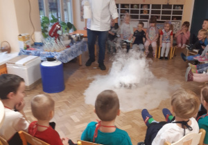 Dzieci siedzą na krzesłach ustawionych w półkole. Na środku na brązowej podłodze widać dym wytwarzany za pomocą suchego lodu.